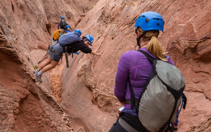 In the foreground, a person looks on as others make their way through a narrow canyon by bracing their hands on one side and their feet on the other. Everyone is wearing helmets.